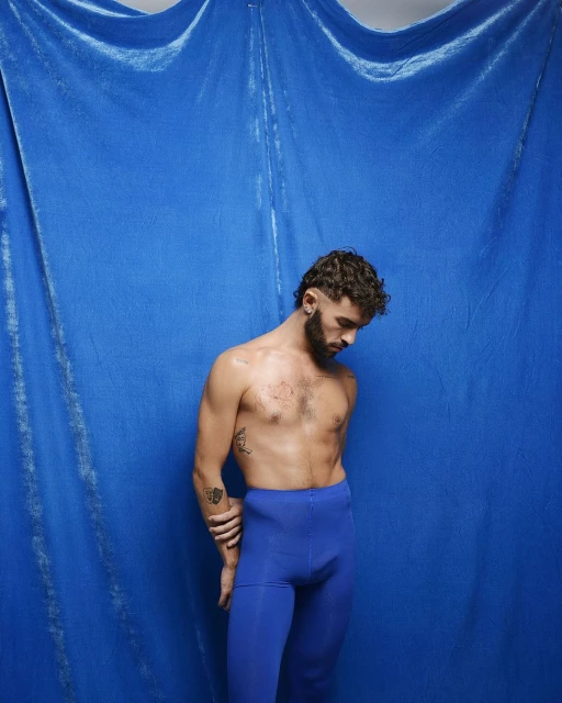 Shirtless male in blue leggings poses against a draped blue velvet backdrop, looking down with curly brown hair and beard in a studio setting.