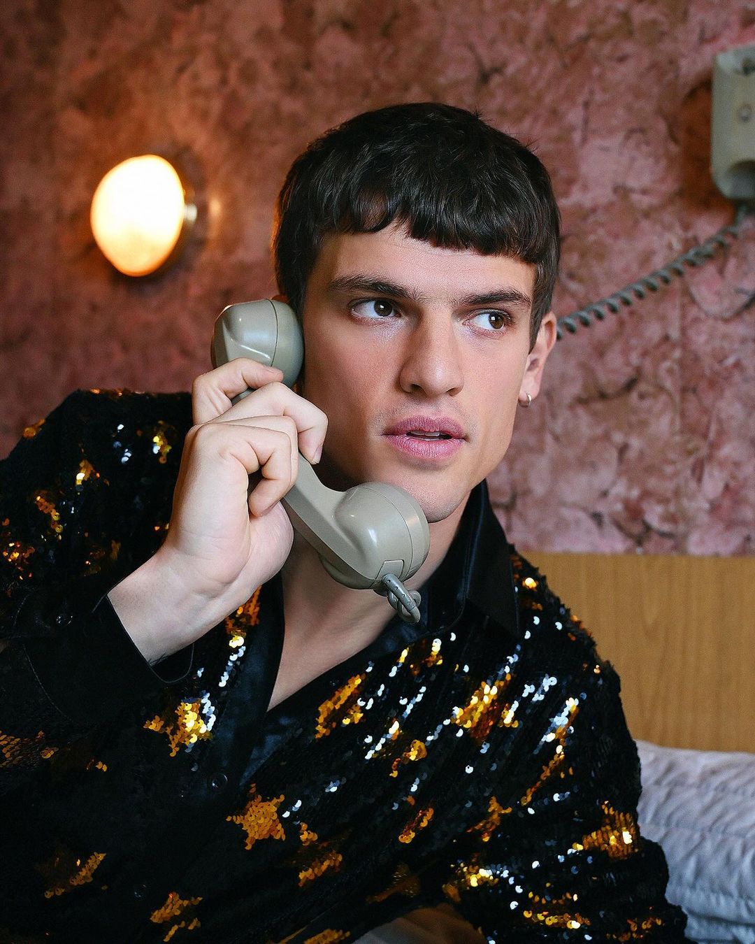 Portrait of a dark-haired adult man wearing a sequin jacket holding a landline telephone receiver in an indoor setting.