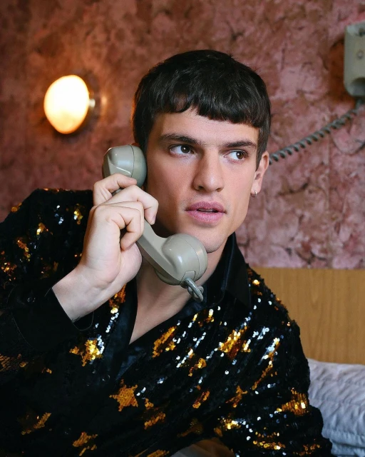 Portrait of a dark-haired adult man wearing a sequin jacket holding a landline telephone receiver in an indoor setting.