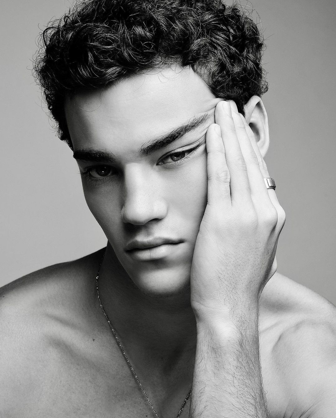 Monochrome portrait of a young adult male with dark, curly hair, gazing intently at the camera with his hand near his face.