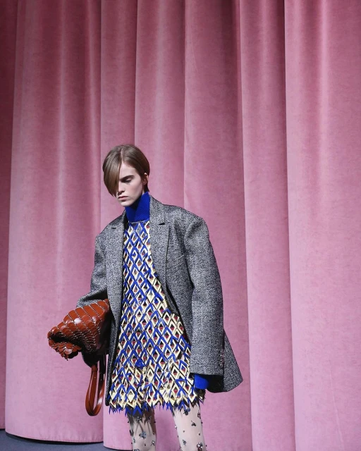 A fashion model poses in a patterned dress and grey jacket, holding a brown purse, against a pink curtain backdrop in a studio.