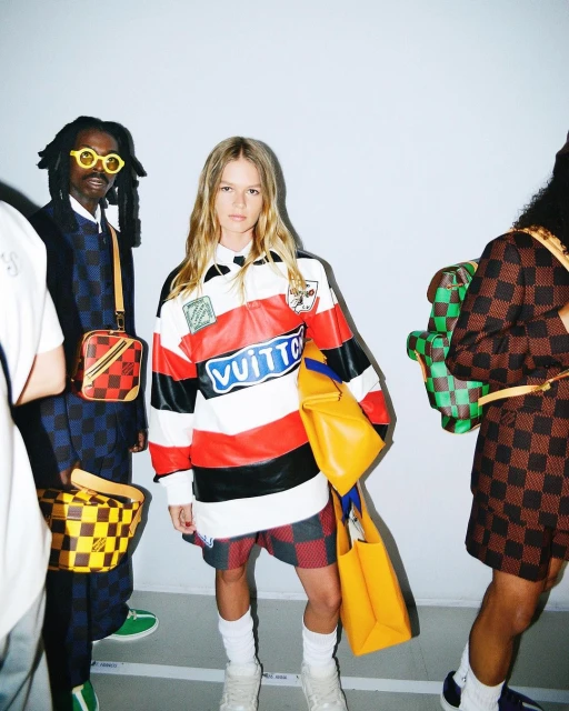 A group of fashionably dressed people pose in front of a plain wall showcasing designer clothing and accessories, with a focus on streetwear.