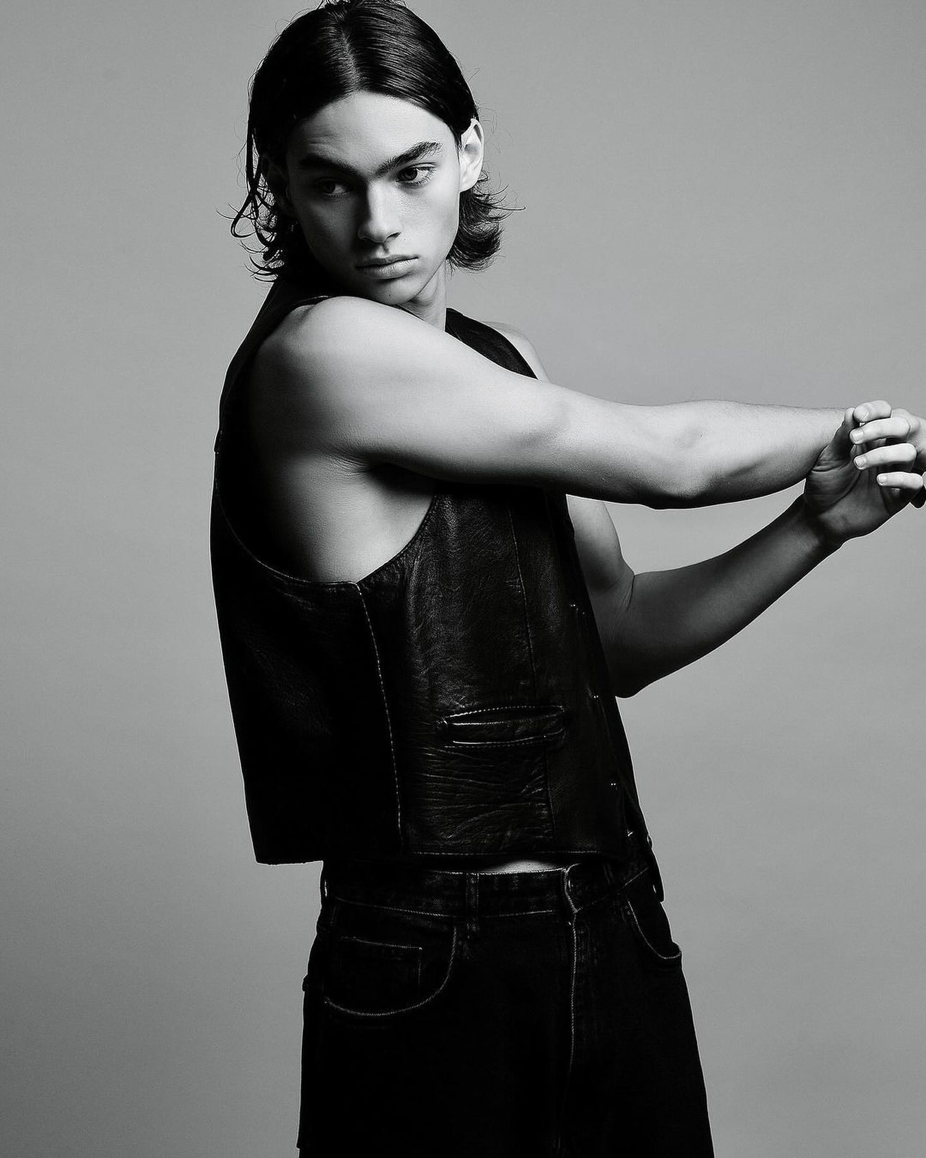 Young male model with dark shoulder-length hair wearing a black leather vest and dark jeans against a grey studio backdrop.