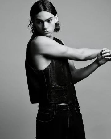 Young male model with dark shoulder-length hair wearing a black leather vest and dark jeans against a grey studio backdrop.