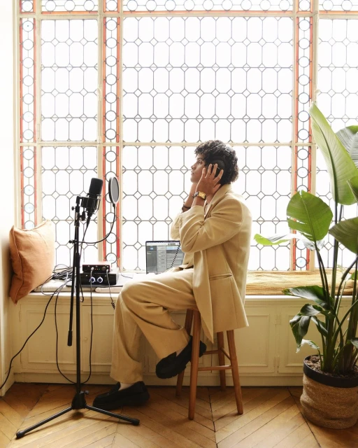 Man in beige suit recording music in a light-filled room with a large window, using a microphone, laptop, and headphones.