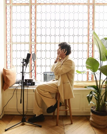Man in beige suit recording music in a light-filled room with a large window, using a microphone, laptop, and headphones.