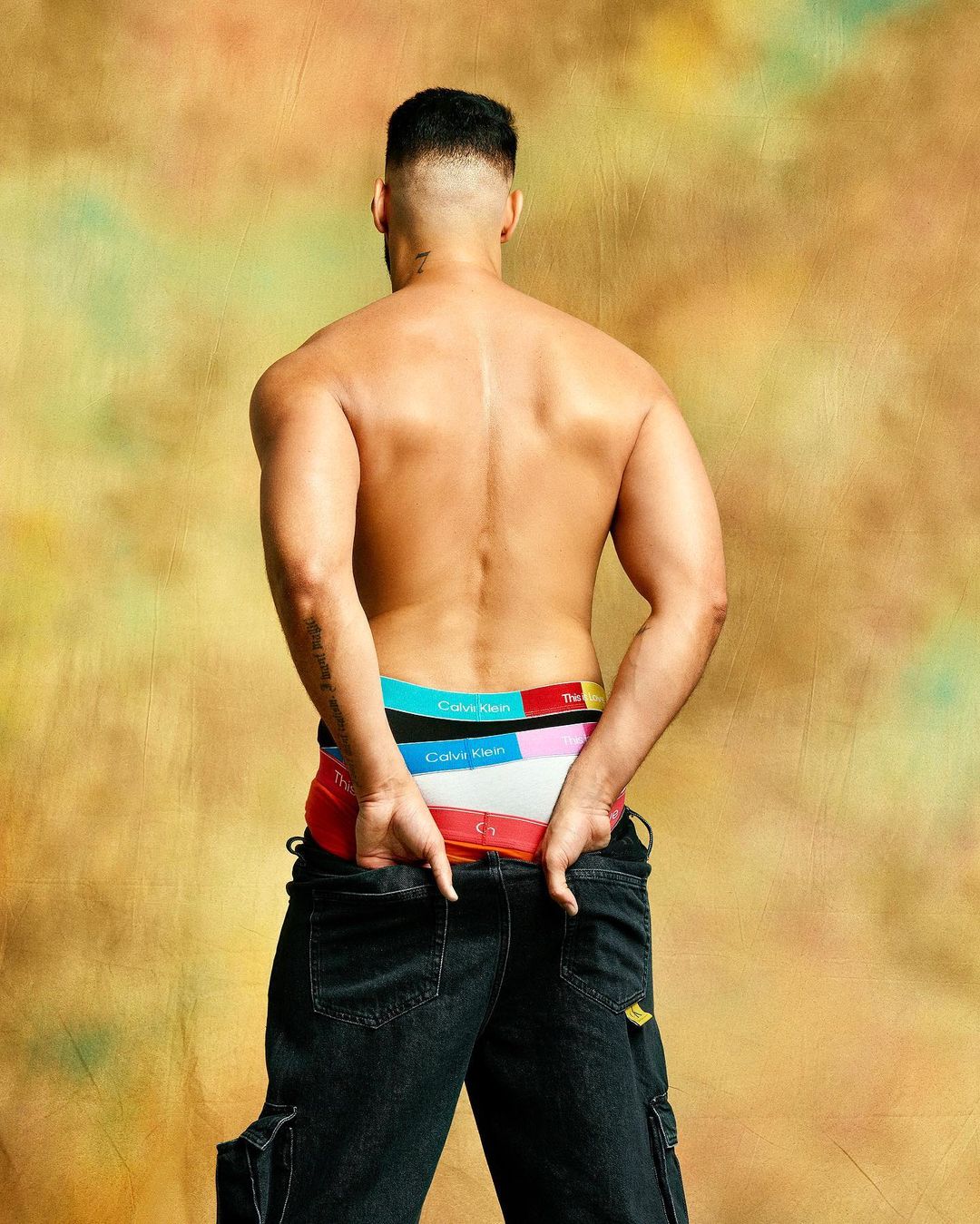 A shirtless male model from the back, wearing colorful Calvin Klein underwear, hands in his denim jeans pockets on a gold mottled backdrop.