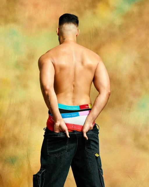 A shirtless male model from the back, wearing colorful Calvin Klein underwear, hands in his denim jeans pockets on a gold mottled backdrop.