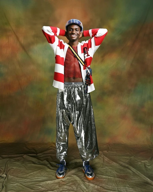 Full-length studio portrait of smiling adult male in shiny silver pants, striped sweater, and cap against mottled neutral backdrop.