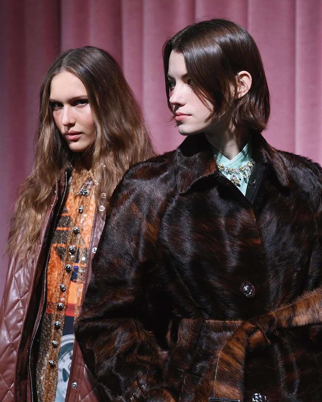 Two fashion models showcase Fall fashion in a studio setting with a brown fur coat and leather jacket against a light pink background.