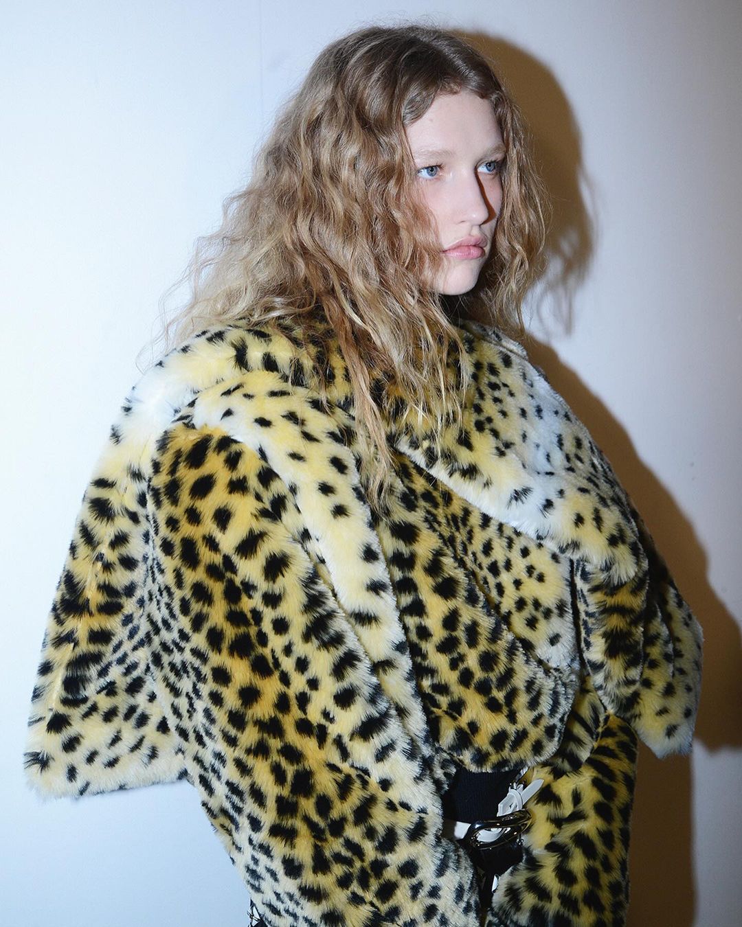 Close-up of a blonde adult female wearing a faux fur coat with a black and yellow leopard print, posing in an indoor environment.