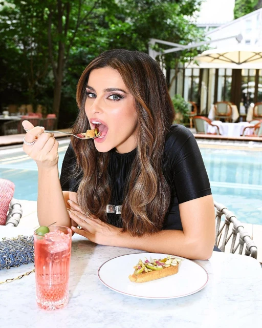 Brunette woman in a black top sits at an outdoor table, eating a snack with a spoon and a pink drink near an outdoor swimming pool.