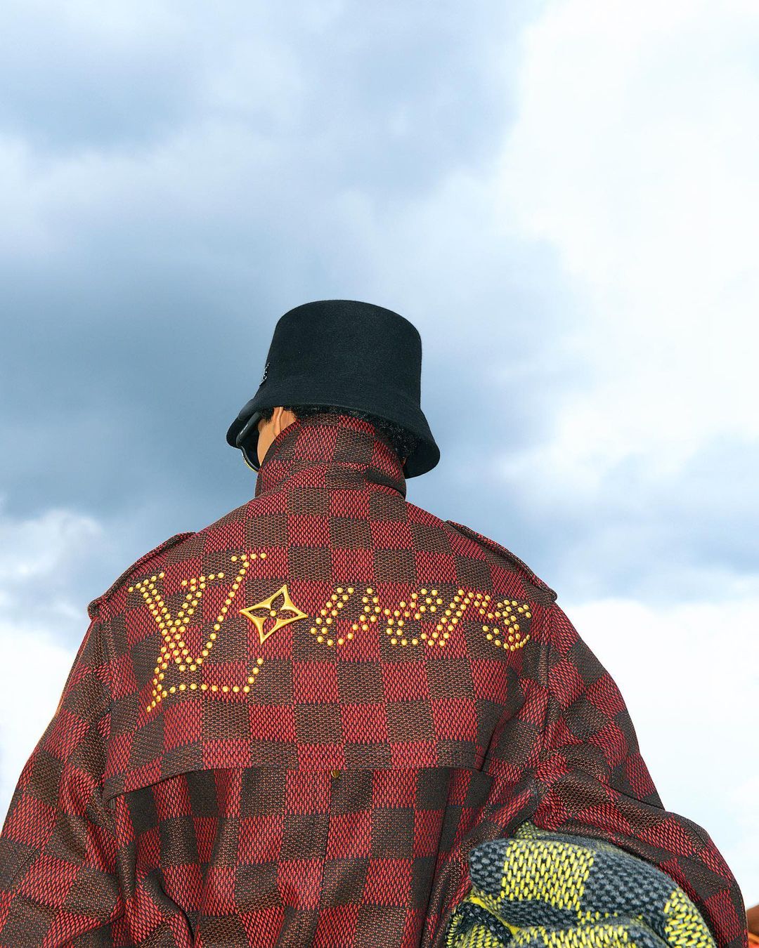 Person wearing Louis Vuitton jacket and black bucket hat against a cloudy sky. The back of the jacket features the LV logo in studs.