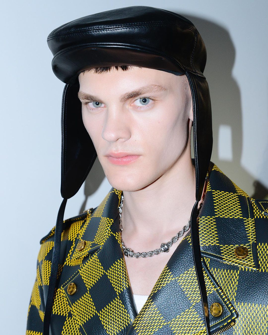 Close-up of a young male model with light blue eyes and wearing a black leather hat and yellow and black jacket in a studio setting.
