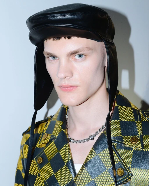 Close-up of a young male model with light blue eyes and wearing a black leather hat and yellow and black jacket in a studio setting.