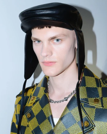 Close-up of a young male model with light blue eyes and wearing a black leather hat and yellow and black jacket in a studio setting.