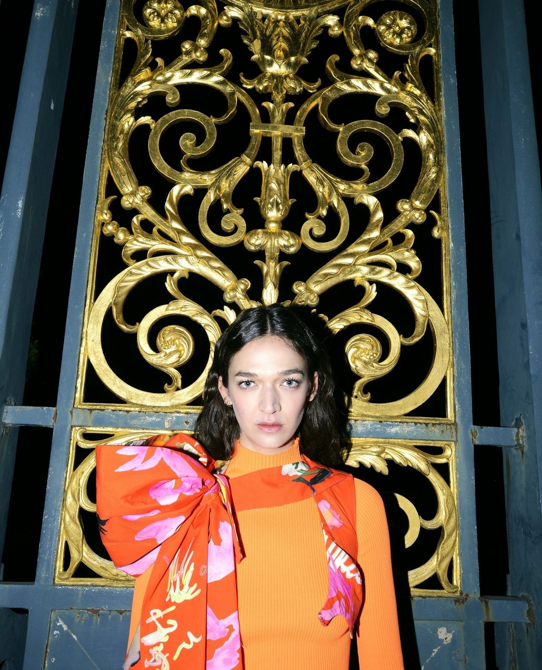 Woman in orange top with floral scarf poses in front of a gold ornamental gate. The gate is set against a black background.