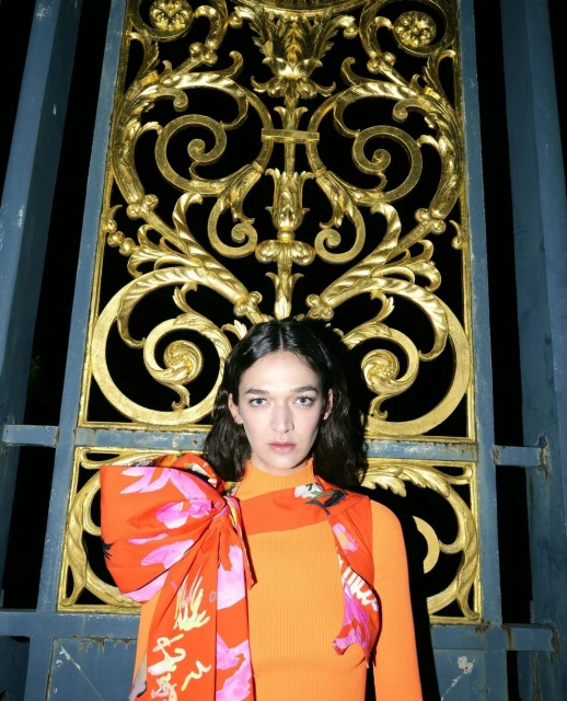 Woman in orange top with floral scarf poses in front of a gold ornamental gate. The gate is set against a black background.