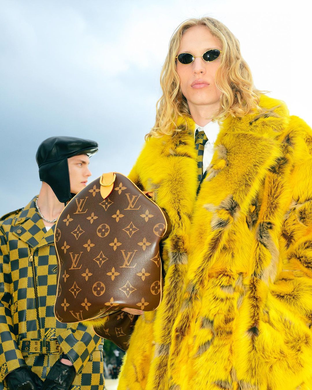 Two men model yellow and checkerboard patterned outerwear with a brown Louis Vuitton handbag against an overcast sky backdrop.