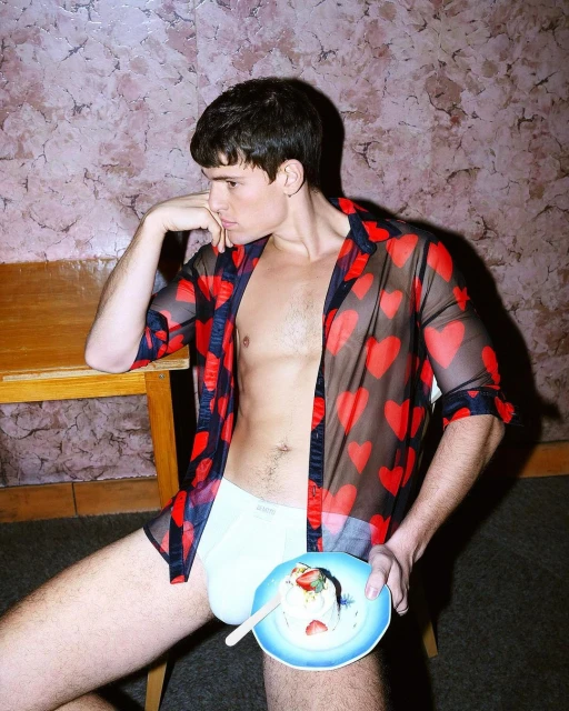 Young adult male poses indoors wearing a sheer heart-print shirt while holding a small dessert plate and sitting near a wooden table.
