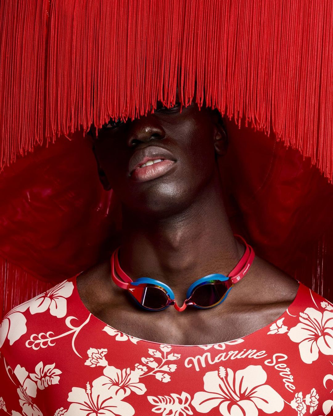 Close-up of a Black male model in a red floral top and swim goggles, framed by a red fringed headdress against a red backdrop.