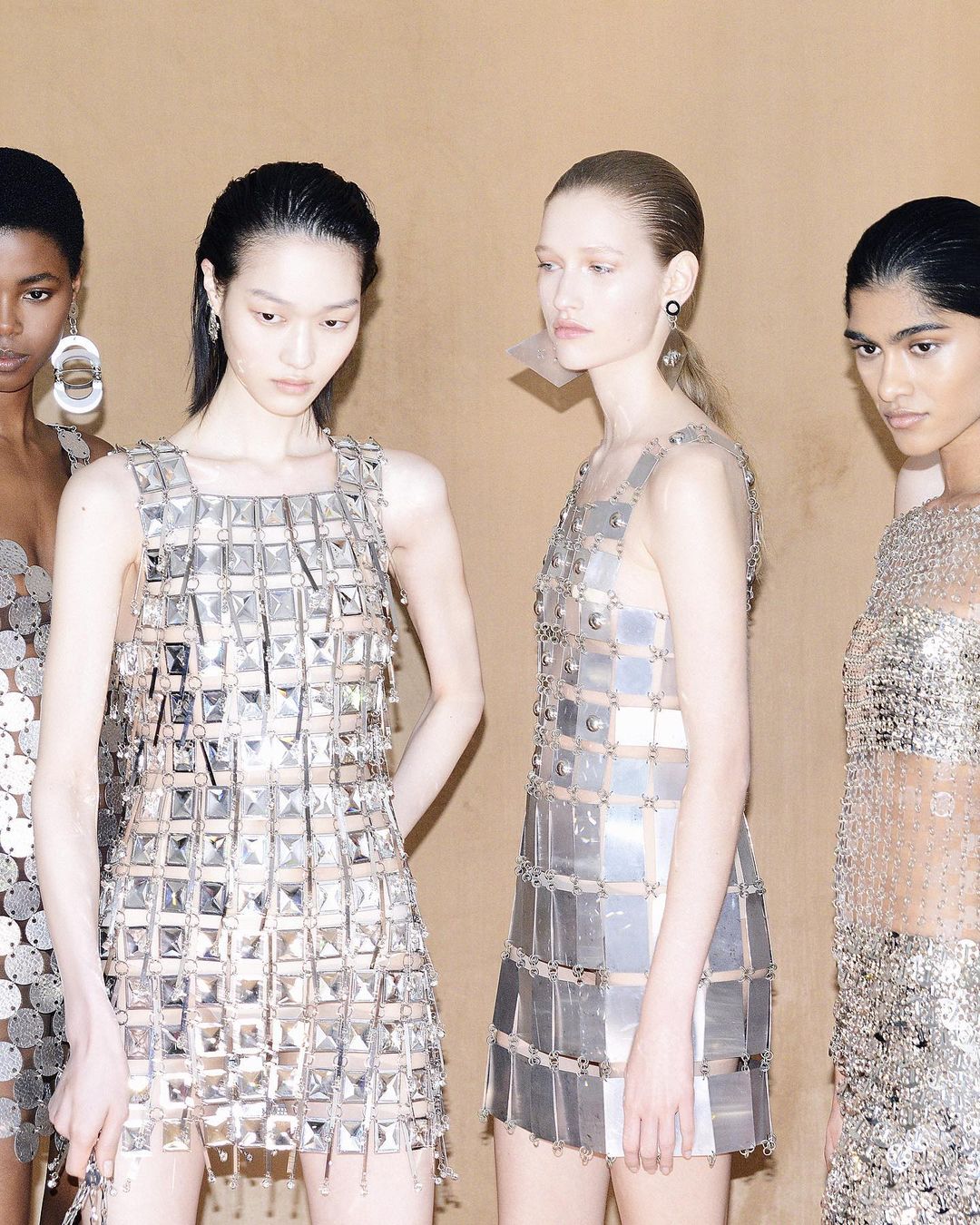 Four women stand shoulder-to-shoulder, wearing reflective silver dresses made of squares and circles in a neutral-toned indoor setting.