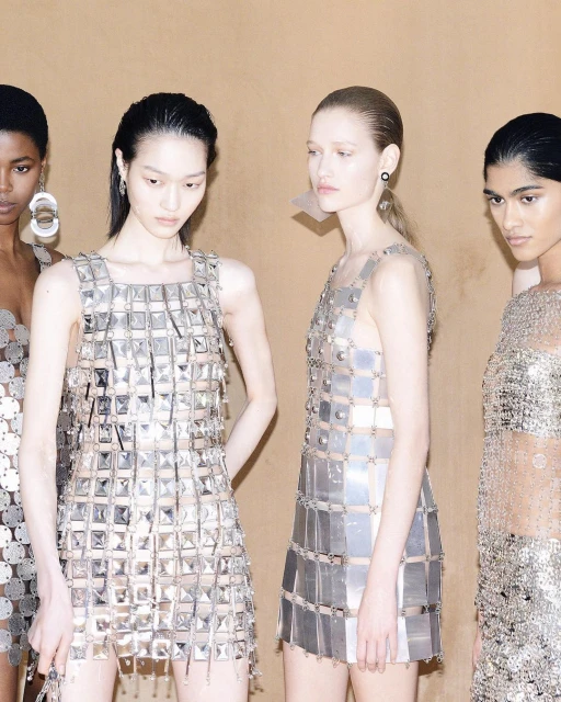 Four women stand shoulder-to-shoulder, wearing reflective silver dresses made of squares and circles in a neutral-toned indoor setting.
