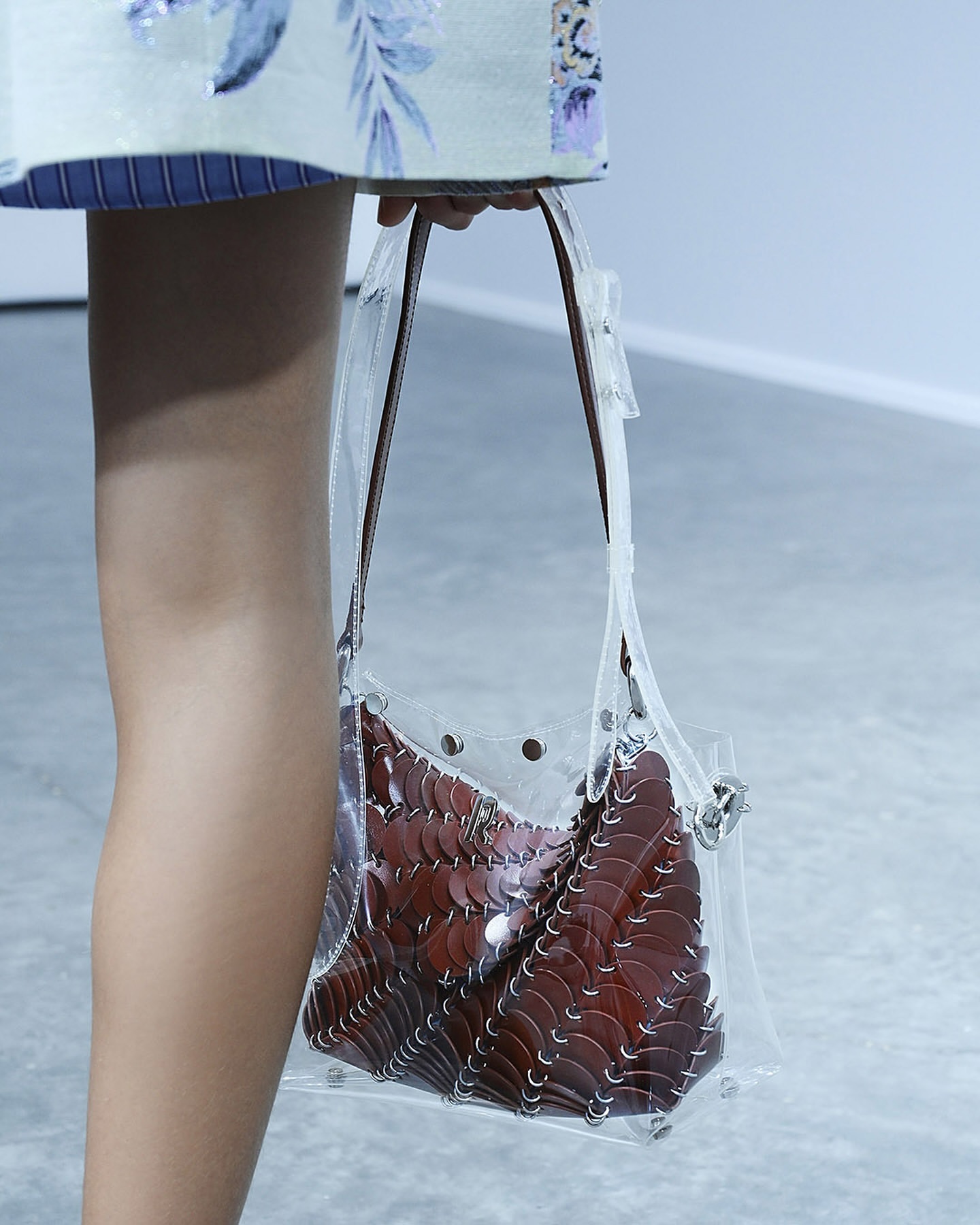 Clear plastic purse with brown scalloped contents, carried by a tan arm and partially viewed person in a light-toned setting.