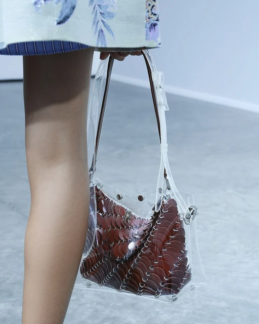 Clear plastic purse with brown scalloped contents, carried by a tan arm and partially viewed person in a light-toned setting.