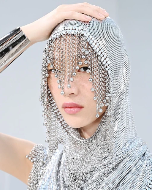 Close-up of an Asian woman wearing a silver metal mesh hooded veil and matching bracelets, posing against a light grey background.