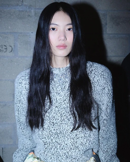 Portrait of an Asian female model with long black hair wearing a speckled grey sweater, posing against a textured grey brick wall.