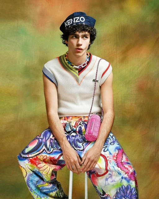 Young adult male in colorful patterned pants sits against a mottled backdrop wearing a Kenzo hat, sleeveless top, and small pink bag.