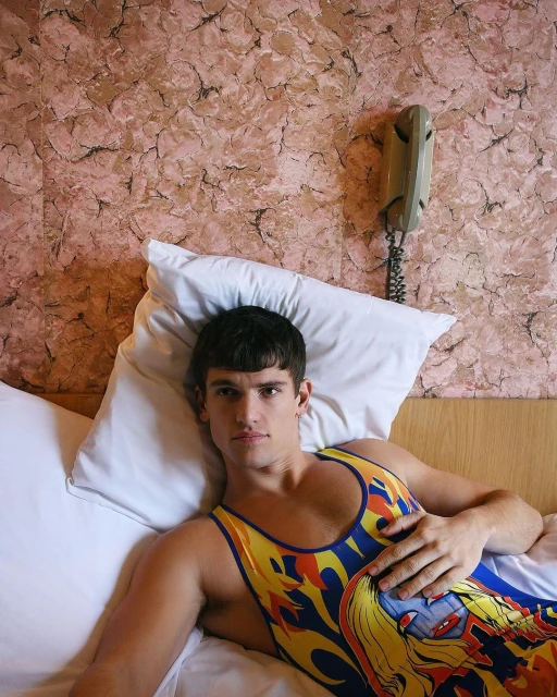 Adult male lying in bed with a graphic print singlet. Pillow and headboard behind him. Landline phone on the textured wall.
