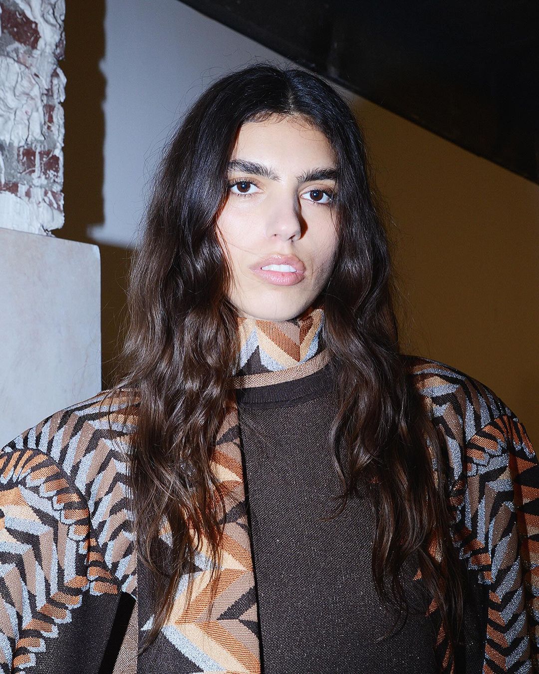 Close-up of an adult woman with long brown hair and fair skin, wearing a patterned brown and tan outfit indoors against a white wall.