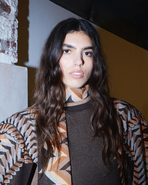 Close-up of an adult woman with long brown hair and fair skin, wearing a patterned brown and tan outfit indoors against a white wall.