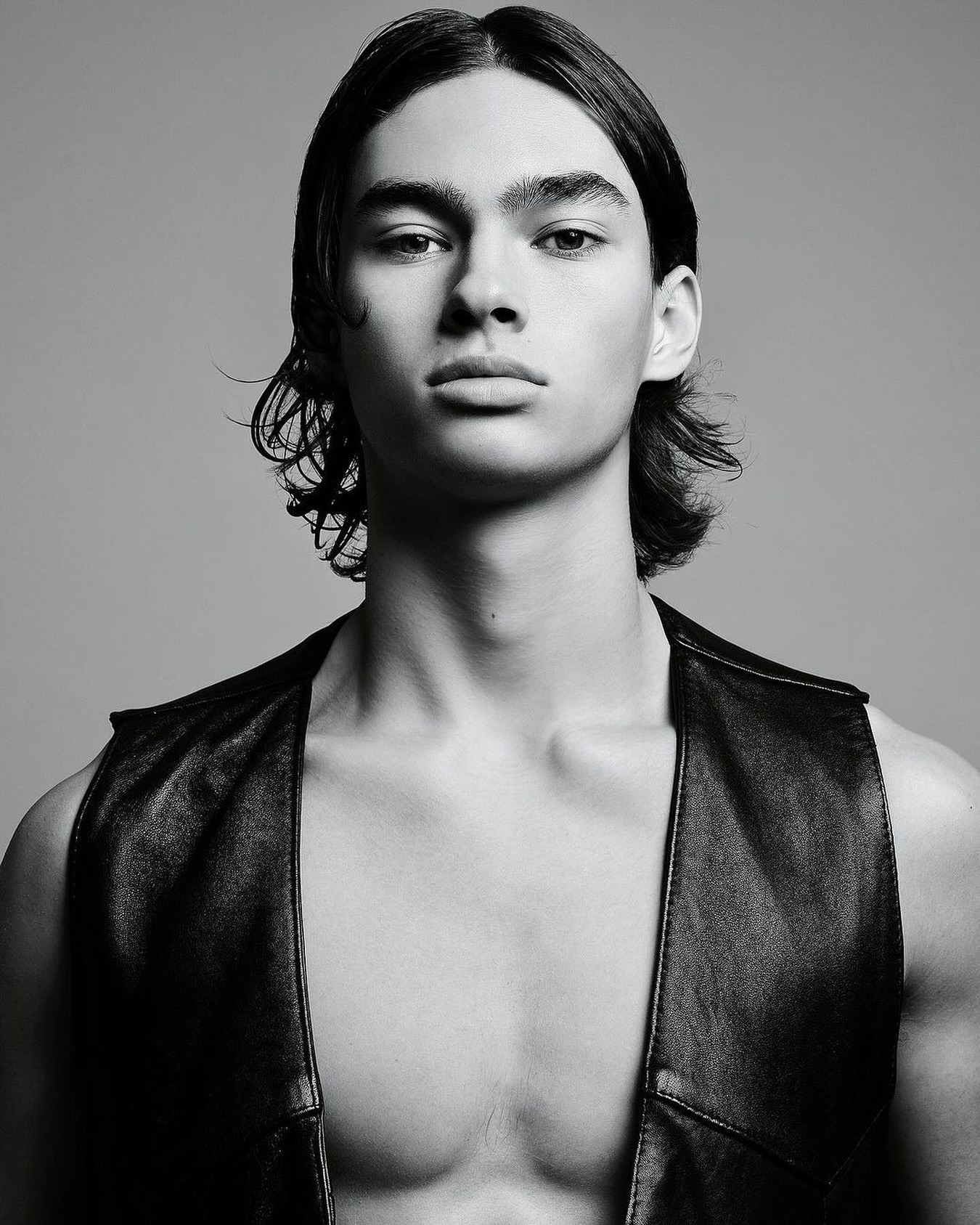 Monochrome portrait of a young adult male model with long hair wearing a leather vest against a neutral gray studio background.