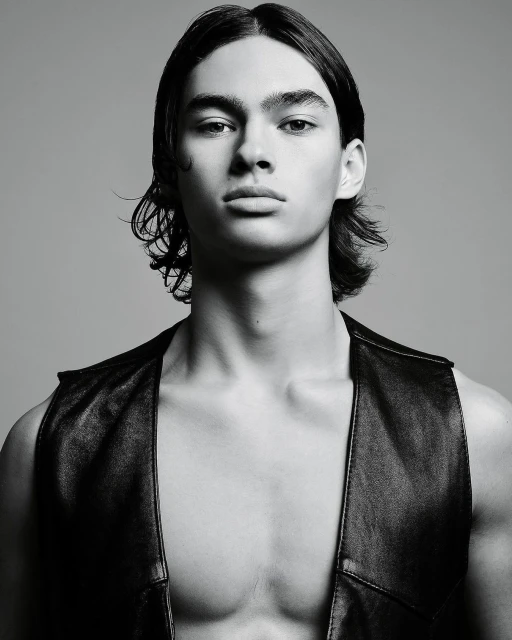 Monochrome portrait of a young adult male model with long hair wearing a leather vest against a neutral gray studio background.