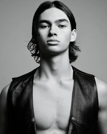 Monochrome portrait of a young adult male model with long hair wearing a leather vest against a neutral gray studio background.