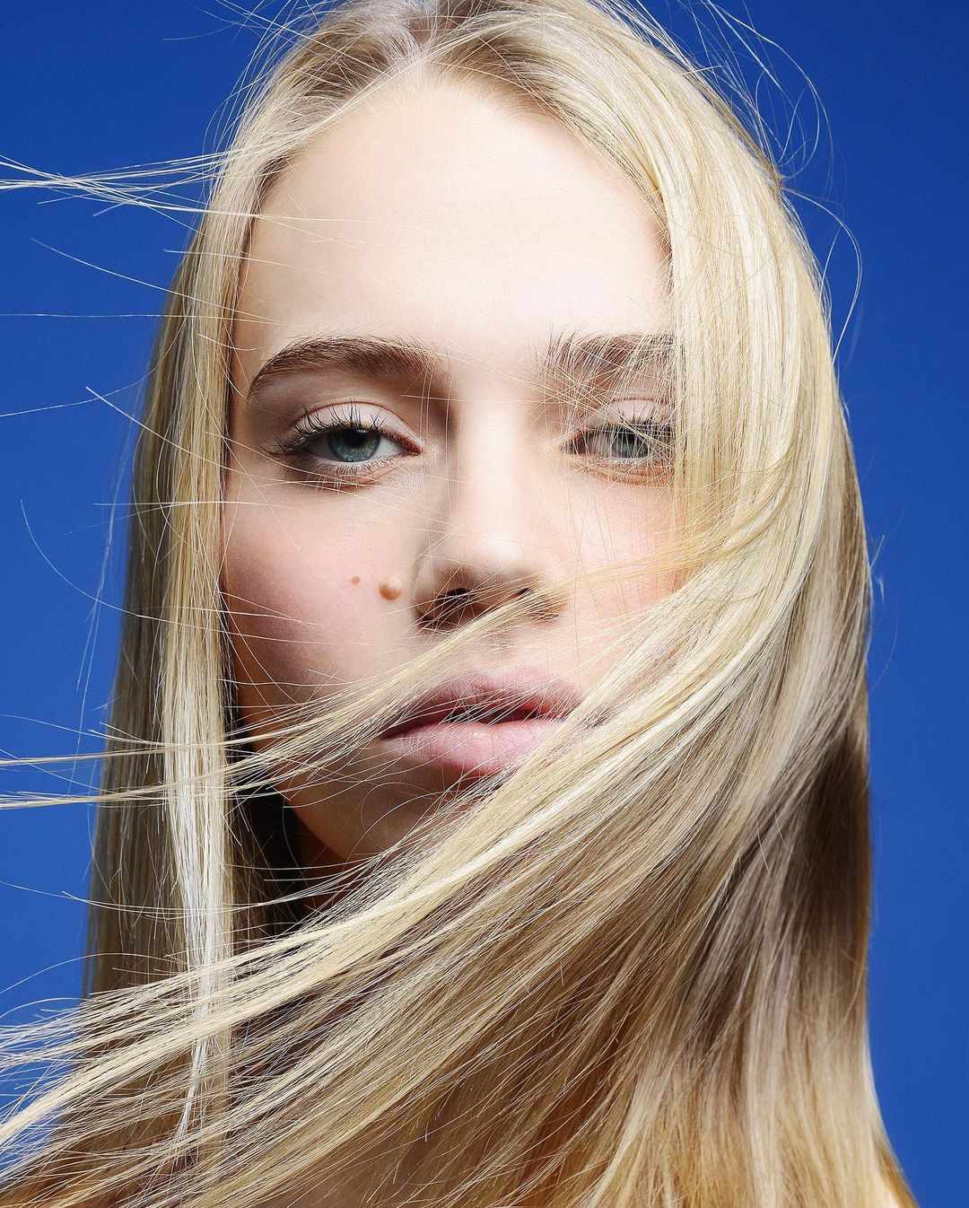 Close-up portrait of a fair-skinned young adult female with blonde hair blowing across her face against a plain blue background.