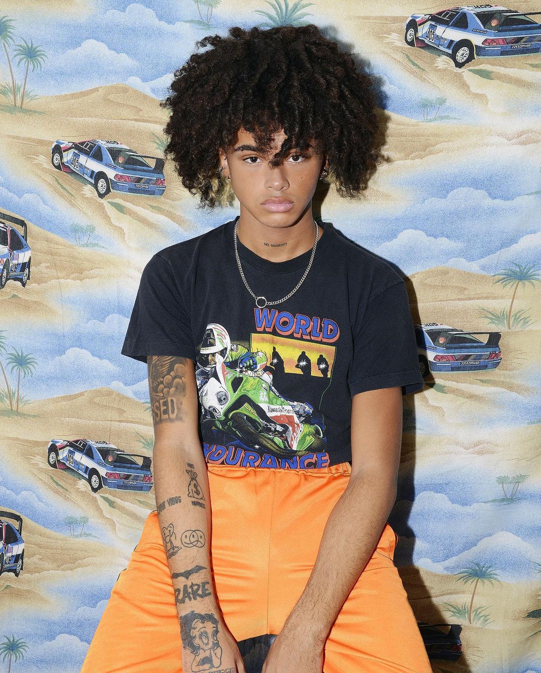 Young adult male poses in orange pants and t-shirt with motorcycle graphic in front of a tropical wallpaper background.