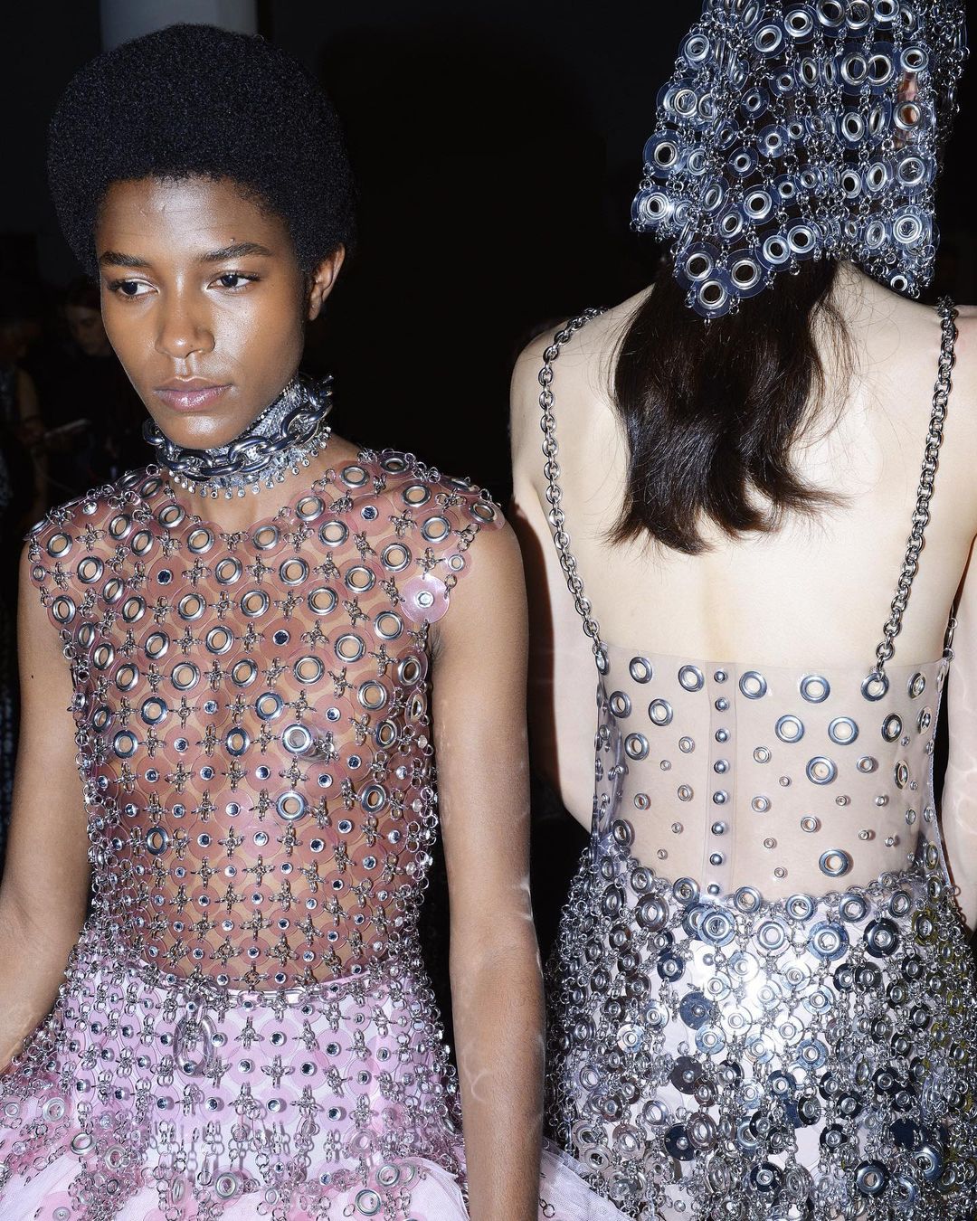 Two women in avant-garde fashion made of metal rings stand in a dark indoor space, presenting unique high-fashion apparel with intricate designs.