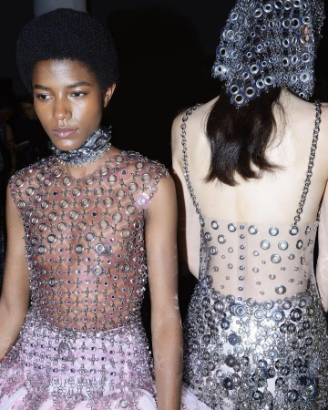 Two women in avant-garde fashion made of metal rings stand in a dark indoor space, presenting unique high-fashion apparel with intricate designs.