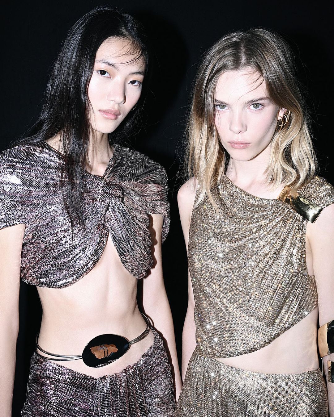 Two fashion models in shimmering metallic outfits pose against a dark backdrop, showcasing asymmetrical tops, stylish accessories, and elegant hair.