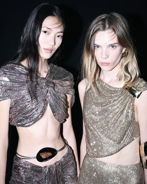 Two fashion models in shimmering metallic outfits pose against a dark backdrop, showcasing asymmetrical tops, stylish accessories, and elegant hair.