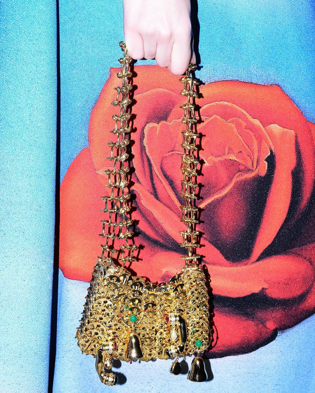 Person holds a gold metal mesh bag with emerald accents against a backdrop featuring a red rose and light blue fabric.