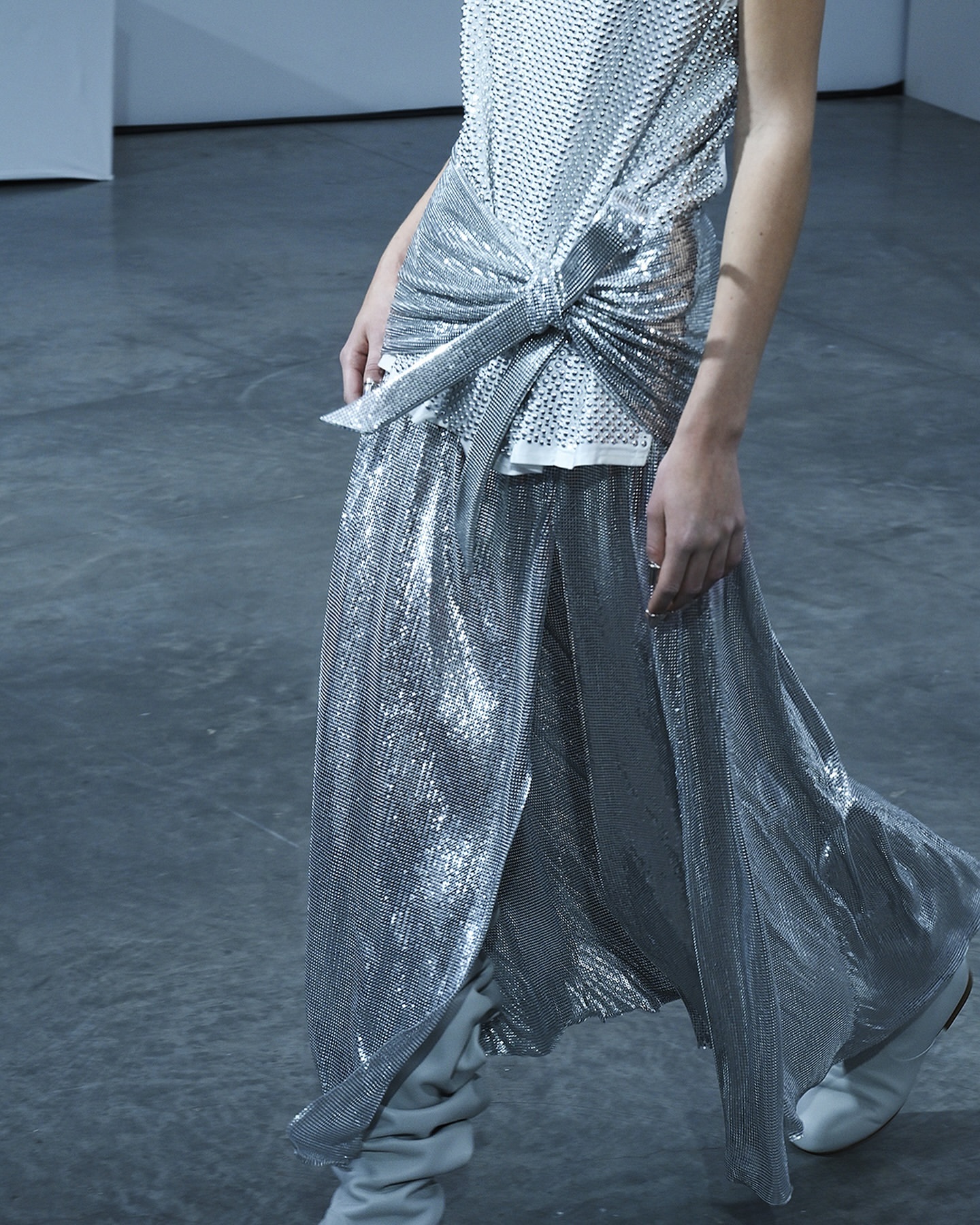 A person is wearing a silver mesh tank top with a knotted front and a matching, high-slit maxi skirt, paired with grey heeled boots on a grey background.