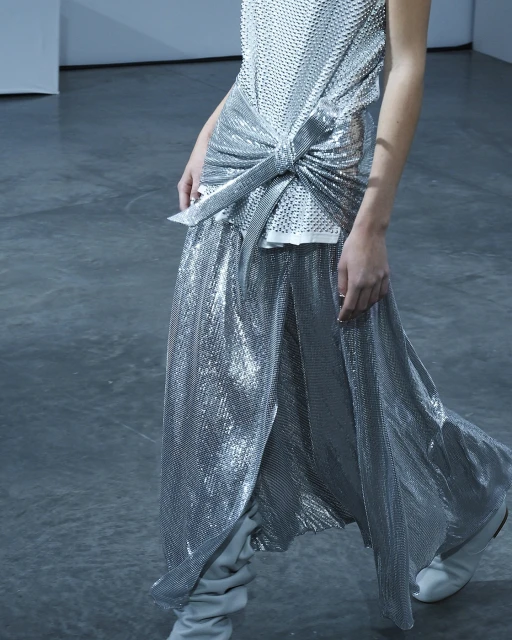 A person is wearing a silver mesh tank top with a knotted front and a matching, high-slit maxi skirt, paired with grey heeled boots on a grey background.