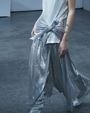 A person is wearing a silver mesh tank top with a knotted front and a matching, high-slit maxi skirt, paired with grey heeled boots on a grey background.