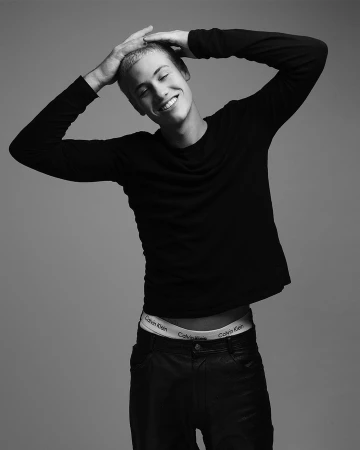 Young man with short hair wearing a black long-sleeve top and trousers against a grey backdrop