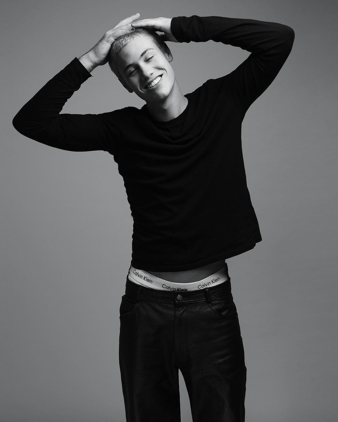 Black and white portrait of a smiling young adult man in dark clothing with his hands on his head in a photo studio setting.